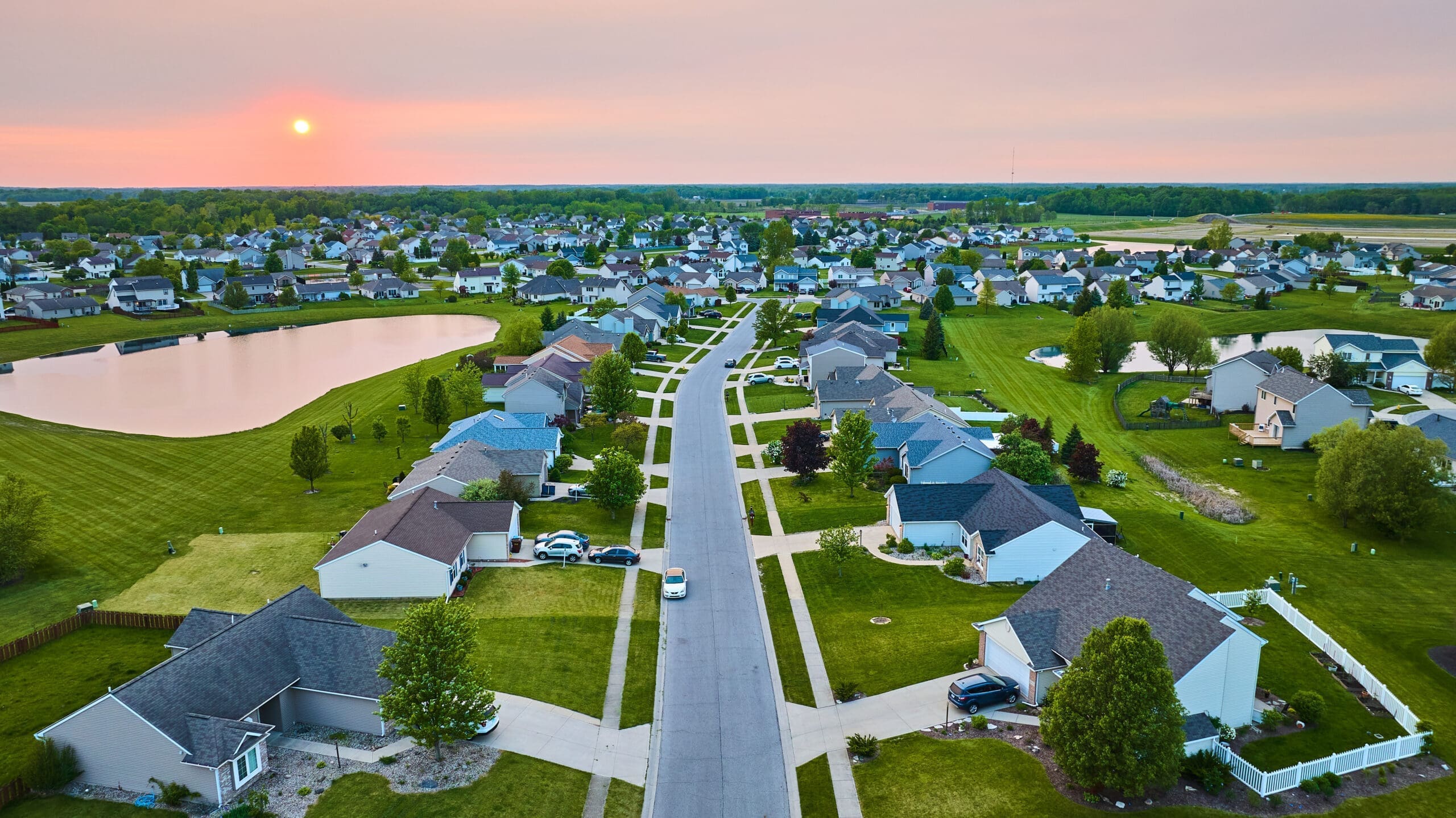 Image of Sunset suburban neighborhood with pond one-story and two-story houses green lawns visible sun aerial
